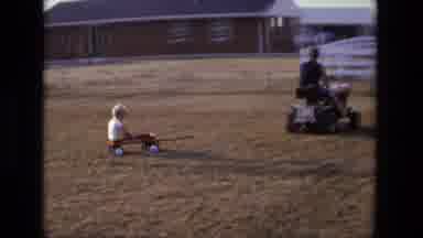 California USA (1975): Kid Riding On Wagon Pulled By Man Driving Lawn Mower In