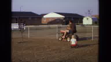 California USA (1975): Man Drives Lawn Mower Around Yard Pulling Kid In Wagon