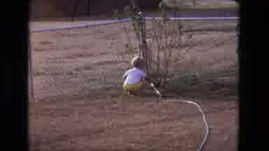 California USA (1975): Young Child In Yellow Shorts And White Shirt Using A Hose