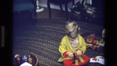 California USA (1975): Young Boy In Yellow Robe Trying To Open A Gift With Toy