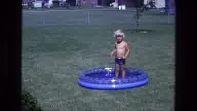 California USA (1975): Boy Wearing Blue Shirt Stands In Blue Kiddie Pool On A