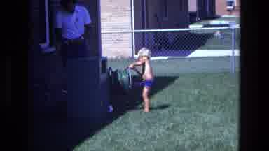 California USA (1975): Child Helps His Father To Collect A Hose Outside His Home