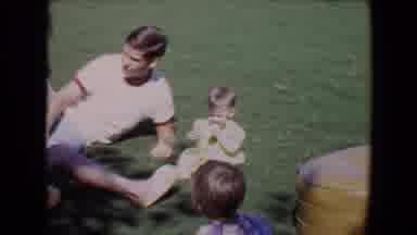 Davis California (1967): A Baby In A Green Dress Sits Outside On Grass As Man Lays