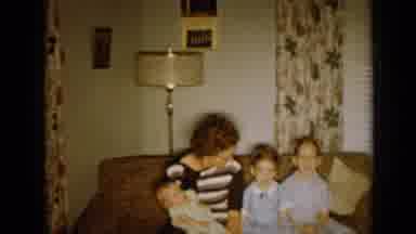 Minnesota USA (1958): Woman Wearing Brown Dress Holds Baby While Sitting On Sofa