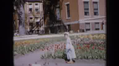 Iowa USA (1959): Girl In Dress And White Hood Walking In The Garden With Many