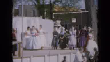 Iowa USA (1959): Woman On A Stage With Patent Dresses On While People Surround The