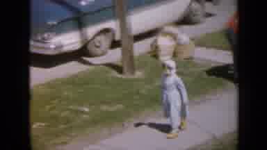 Iowa USA (1959): Child Runs Along Sidewalk Under The Eyes Of His Parents On A