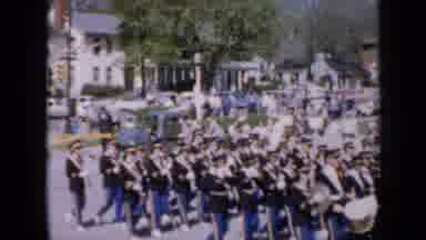 Iowa USA (1959): Marching Band In Festival Marching Down Road As Crowd Watches