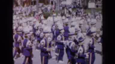 Iowa USA (1959): Marching Band In Festival Marching Down Road As Crowd Watches
