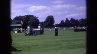 England (1963): Black Dog Runs To Men Walking On Grass On Sunny Day