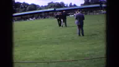 England (1963): Man Fires Starter Gun At Person Wearing Black Coat On A Sunny Day