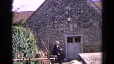 France (1963): Man Wearing Black Jacket Walks Past Stone Building On A Bright