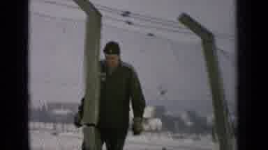 Germany Military Base (1963): Man Wearing Grey Jacket And Hat Walks Past Chain