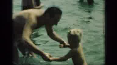 New York (1949): Man Wearing Tan Shorts Holds Hands Of Toddler In Water On A