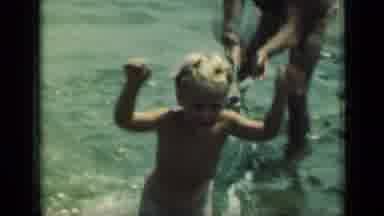 New York (1949): Man Wearing Tan Shorts Helps Small Boy Stand In Water On A Bright