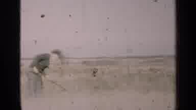 Illinois USA (1947): Man Wearing Black Shirt Uses Scythe Near White Building On A