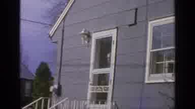 Long Island New York (1965): Woman Wearing Yellow Coat Stands Next To Gray House