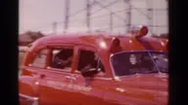 Mineola New York (1966): People In A Red Car Drive Through A Busy Street By