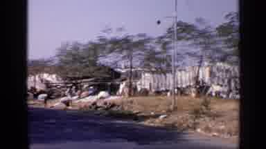 India (1970): A Shanty Town Beside A Street As A Car Drives By Filming