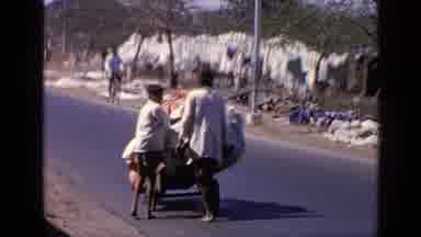 India (1970): Two Men Push Wares Down Street As Bike Rider Moves By Them