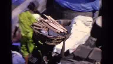 India (1970): Boy Carrying Wood On His Head In A Basket While Another Boy Washes