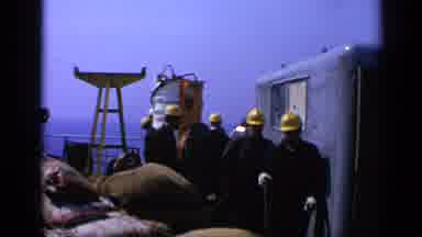 United Kingdom (1970): Workers In Dark Outfits And Yellow Hard Hats File By Bags