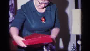 California USA (1969): A Woman In A Blue Dress Wearing Glasses Opens A Gift From A