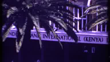 Kenya (1969): Storefront Behind Palm Trees And Busy Street And Parking Lot Filmed