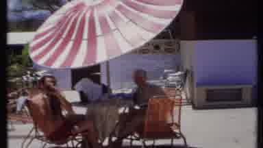 California USA (1971): People Sit In Lawn Chairs At A Table Beneath A Red And