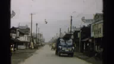 Japan (1952): Vehicles Drive On A Road Between Houses While People Pedal Bicycles