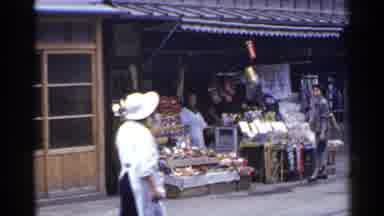 Japan (1953): Woman Wearing Gray Suit Walks Past Produce Stand On A Clear Day