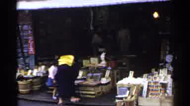 Japan (1953): Woman Wearing Tan Dress Looks At Items Of Outdoor Market On A Clear