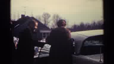Ohio USA (1959): Man Wearing Black Coat Helps Woman Get Into White Car On A Sunny