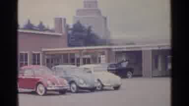 Germany (1961): Three Volkswagen Beetles Parked By Brown Building On A Clear Day