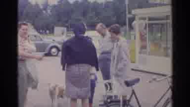 Germany (1961): Adults And Dog Stand Around Child Holding Box In Parking Lot Near
