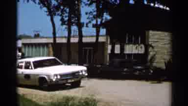 Hamel Minnesota (1966): White Car Parked By Brown Building On A Bright Sunny Day