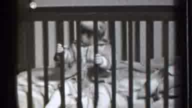 Danielson Connecticut (1939): Baby Wearing Grey Coat Sits In Wooden Crib Holding