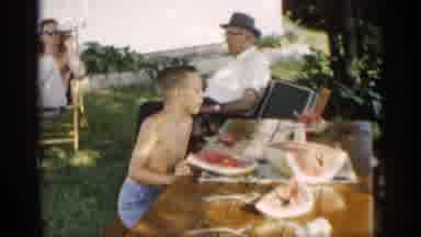 Indiana (1959): Kid Eating Watermelon And Family Playing With Dogs Behind Fence