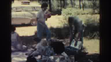 Rhodes Greece (1962): Woman Wearing White Shirt Sets Red Object Down Near Picnic