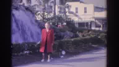 Florida USA (1962): Woman Wearing Red Coat Stands Next To Waterfall On A Clear Day