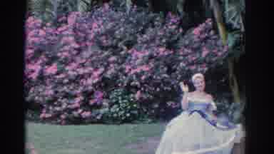 Florida USA (1962): Woman Wearing White Dress Sits On Tan Bench Waving At Camera