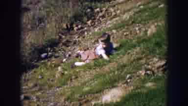 Nebraska (1956): Toddler Wearing Brown Pants Climbs Up Green Hill On A Sunny Day
