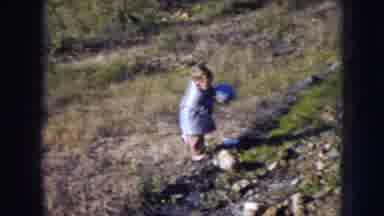 Nebraska (1956): Toddler Wearing Blue Coat Carries Blue Ball Up Green Hill On A