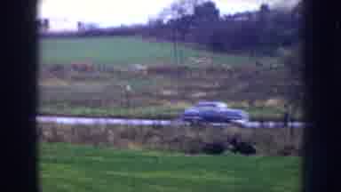 Nebraska (1956): Blue Car Speeds By Pale Yellow House On A Clear Day
