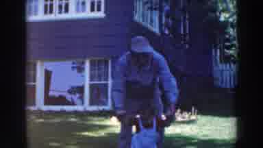 Tustin Wisconsin (1964): Man Wearing Tan Hat Helps Baby Wearing Red Shirt To Walk