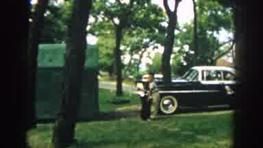 Wisconsin (1957): Boy Wearing Tan Pants Stands Next To Parked Black Car On A Clear