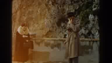 Italy (1960): Man Wearing Black Hat Picks Up Woman Wearing Tan Coat On A Clear Day