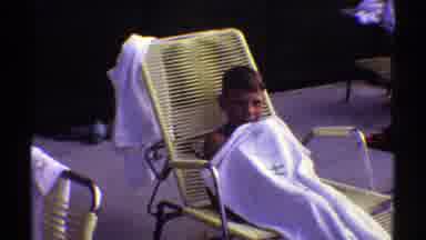 Arizona (1969): Boy Sits In Yellow Chair Holding White Towel And Waving At Camera
