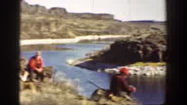 California (1936): Two Men Wearing Red Shirts Sit At River Edge Aiming Rifles At