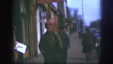 Kansas (1958): Two Men Wearing Grey Shirts Walk Past Red Building On A Clear Day